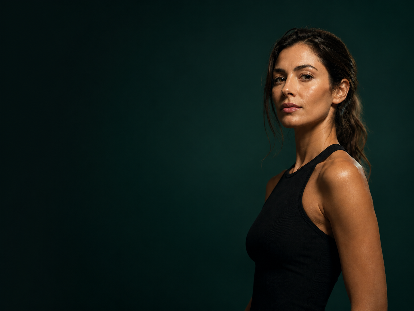 A woman in a black top standing against a deep forest-green background, looking toward the camera